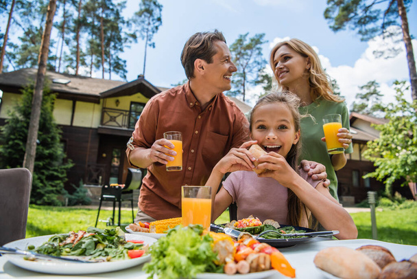Positive kid looking at camera near food and parents outdoors  - Photo, image
