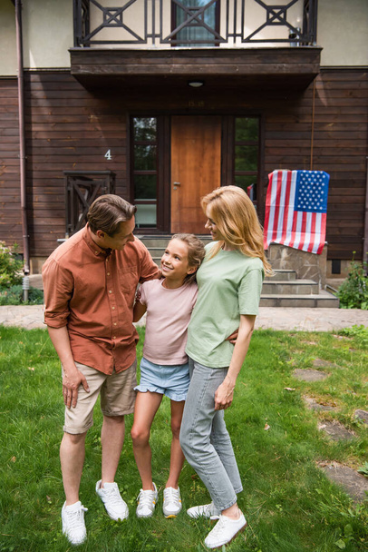 Smiling parents hugging daughter near blurred american flag on vacation house  - Fotó, kép