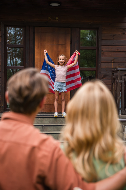 Enfant joyeux tenant drapeau américain près des parents flous et de la maison de vacances  - Photo, image