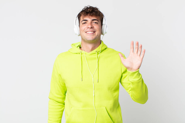 young handsome man smiling happily, waving hand, welcoming and greeting you and headphones - 写真・画像