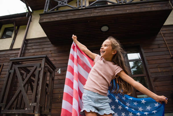 Visão de ângulo baixo de menina sorridente segurando bandeira americana perto da casa  - Foto, Imagem