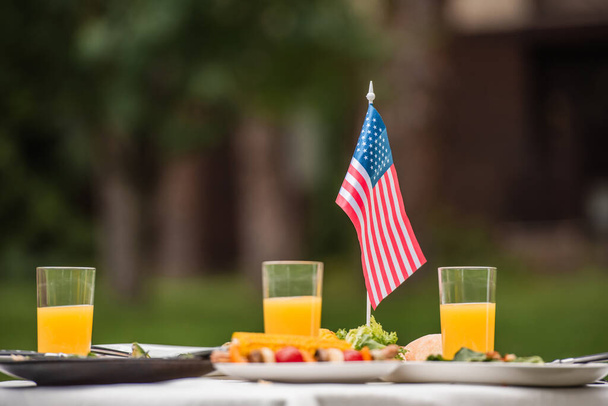 Bandera americana cerca de zumo de naranja y alimentos desenfocados al aire libre  - Foto, Imagen