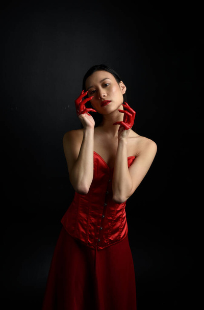 Nahaufnahme Porträt einer schönen jungen asiatischen Frau, die ein rotes Korsett trägt, mit blutiger Farbe überzogen und im gotischen Horrorstil posiert, mit kreativen Handgesten isoliert vor dunkelschwarzem Studiohintergrund. - Foto, Bild