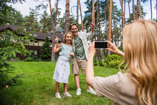 Sonriente padre e hijo sosteniendo raquetas de bádminton cerca de la madre tomando fotos en el teléfono inteligente al aire libre  - Foto, Imagen