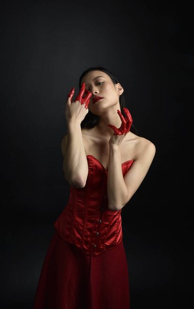 Close up portrait of beautiful young asian woman wearing red corset, covered with bloody  paint posing in gothic horror style with creative hand gestures isolated against dark black studio background. - 写真・画像