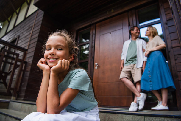 Girl looking at camera on stairs near blurred parents and vacation house  - Foto, afbeelding