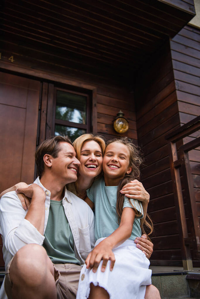 Família sorridente com olhos fechados abraçando no alpendre da casa de férias  - Foto, Imagem