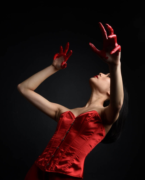 Close up portrait of beautiful young asian woman wearing red corset, covered with bloody  paint posing in gothic horror style with creative hand gestures isolated against dark black studio background. - Фото, зображення