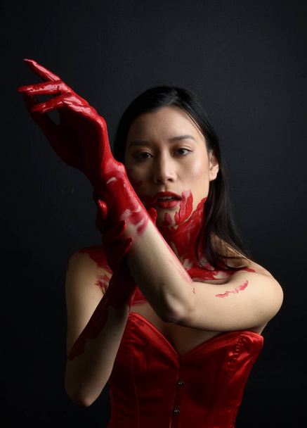 Kırmızı korse giyen güzel Asyalı bir kadının yakın plan portresi, kanlı boyayla kaplanmış, Gotik korku tarzında, yaratıcı el hareketleriyle koyu siyah stüdyo arka planına izole edilmiş.. - Fotoğraf, Görsel