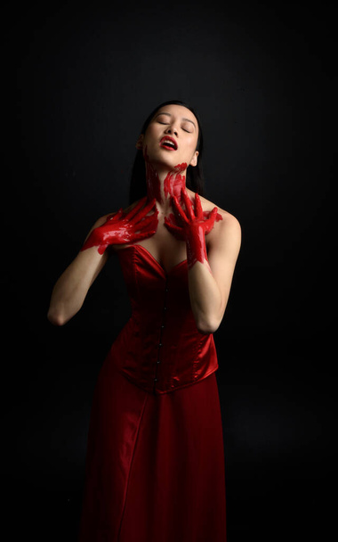 Close up portrait of beautiful young asian woman wearing red corset, covered with bloody  paint posing in gothic horror style with creative hand gestures isolated against dark black studio background. - Foto, Imagen