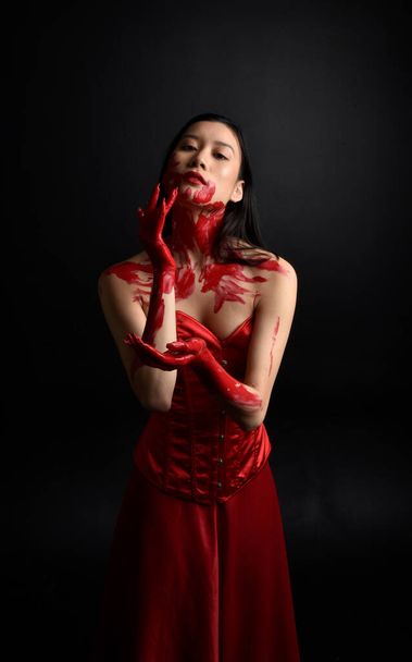 Close up portrait of beautiful young asian woman wearing red corset, covered with bloody  paint posing in gothic horror style with creative hand gestures isolated against dark black studio background. - Valokuva, kuva