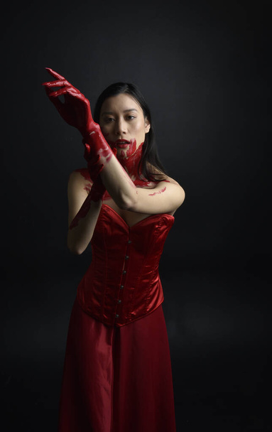 Close up portrait of beautiful young asian woman wearing red corset, covered with bloody  paint posing in gothic horror style with creative hand gestures isolated against dark black studio background. - Fotografie, Obrázek