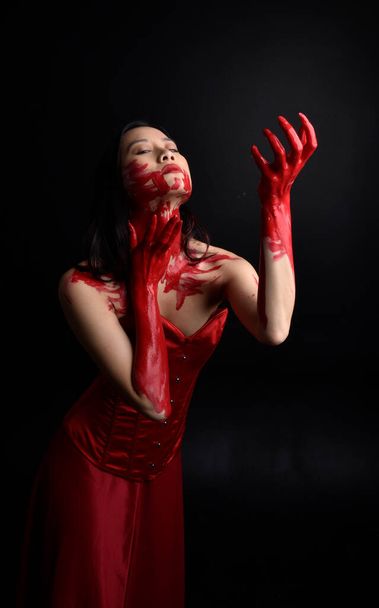Close up portrait of beautiful young asian woman wearing red corset, covered with bloody  paint posing in gothic horror style with creative hand gestures isolated against dark black studio background. - Foto, Bild