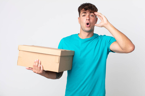 young handsome man looking happy, astonished and surprised and holding a cardboard box - Fotó, kép