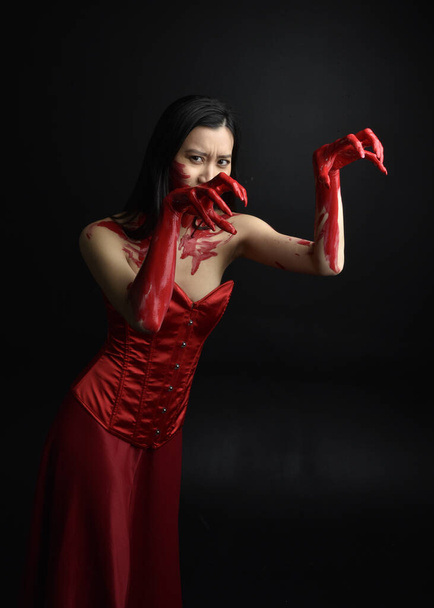 Közelkép portré gyönyörű fiatal ázsiai nő visel piros fűző, borított véres festék pózol gótikus horror stílusban kreatív kéz gesztusok elszigetelt ellen sötét fekete stúdió háttér. - Fotó, kép