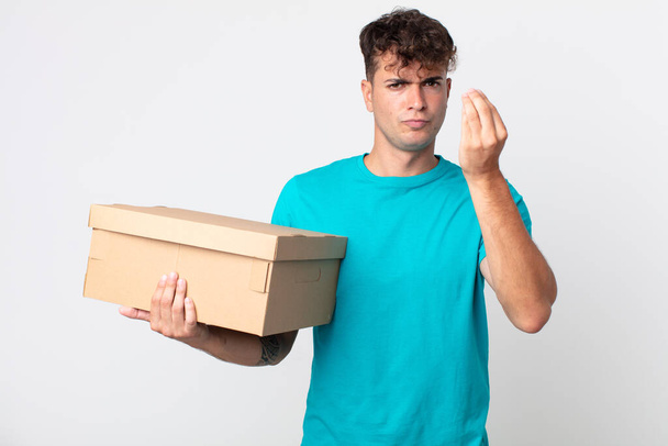 young handsome man making capice or money gesture, telling you to pay and holding a cardboard box - Foto, Imagen