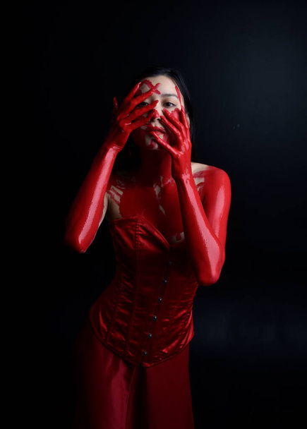 Крупным планом портрет красивой молодой азиатской женщины в красном корсете, покрытый окровавленной краской, позирующей в готическом стиле ужасов, с творческими жестикуляциями рук, изолированными на темном черном фоне студии. - Фото, изображение