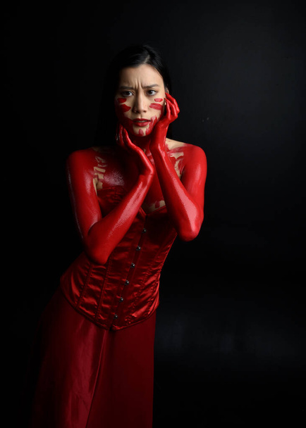 Close up portrait of beautiful young asian woman wearing red corset, covered with bloody  paint posing in gothic horror style with creative hand gestures isolated against dark black studio background. - Foto, imagen