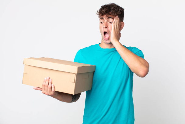 young handsome man feeling happy, excited and surprised and holding a cardboard box - Fotó, kép