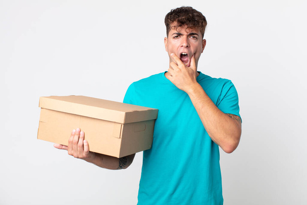young handsome man with mouth and eyes wide open and hand on chin and holding a cardboard box - Fotoğraf, Görsel
