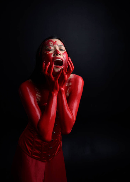 close-up portret van mooie jonge aziatische vrouw dragen rood korset, bedekt met bloederige verf poseren in gotische horror stijl met creatieve hand gebaren geïsoleerd tegen donker zwart studio achtergrond. - Foto, afbeelding