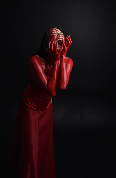 Κοντινό πορτραίτο όμορφης νεαρής Ασιάτισσας με κόκκινο κορσέ, καλυμμένο με ματωμένη μπογιά που ποζάρει σε γοτθικό ύφος τρόμου με δημιουργικές χειρονομίες απομονωμένες σε σκούρο μαύρο φόντο στούντιο. - Φωτογραφία, εικόνα