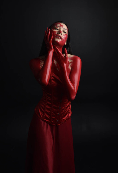 Close up portrait of beautiful young asian woman wearing red corset, covered with bloody  paint posing in gothic horror style with creative hand gestures isolated against dark black studio background. - Photo, Image