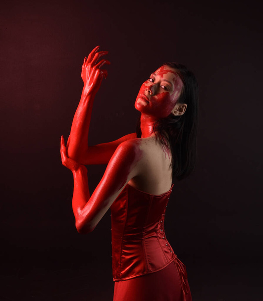 Close up portrait of beautiful young asian woman wearing red corset, covered with bloody  paint posing in gothic horror style with creative hand gestures isolated against dark black studio background. - Zdjęcie, obraz