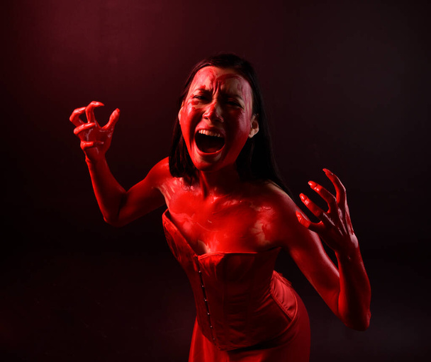 close-up portret van mooie jonge aziatische vrouw dragen rood korset, bedekt met bloederige verf poseren in gotische horror stijl met creatieve hand gebaren geïsoleerd tegen donker zwart studio achtergrond. - Foto, afbeelding