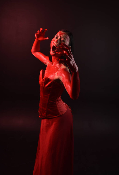Close up portrait of beautiful young asian woman wearing red corset, covered with bloody  paint posing in gothic horror style with creative hand gestures isolated against dark black studio background. - Foto, Imagen