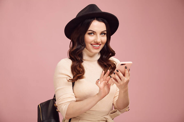黒の帽子のかわいい笑顔ブルネットの女性、スタイリッシュな服を着て彼女の電話を使用し、ピンクの隔離されたカメラを見て. - 写真・画像