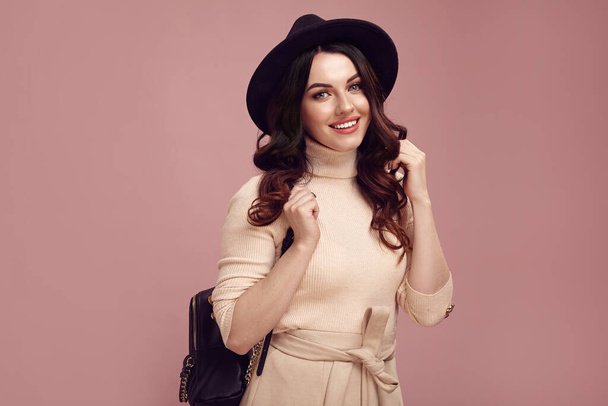 Очаровательная леди с милым взглядом одета в стильную повседневную одежду, носит черную шляпу и рюкзак, искренне улыбаясь против розового изолированы - Фото, изображение
