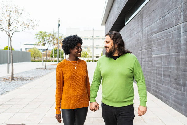 Joven pareja multirracial sonriente cogida de la mano mientras camina por la calle. Hombre y mujer enamorados caminando afuera. - Foto, Imagen