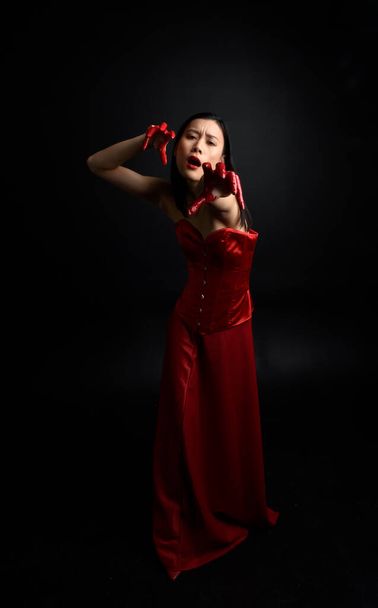 Full length portrait of beautiful young asian woman wearing red corset, covered with bloody paint posing in gothic horror style with creative hand gestures isolated against dark black studio background. - Zdjęcie, obraz