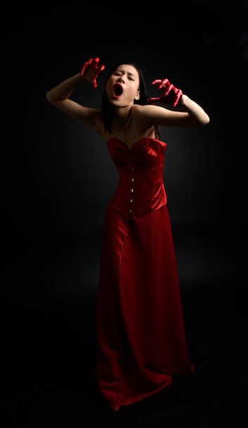 赤のコルセットを身に着けている美しい若いアジアの女性の完全な長さの肖像画,暗い黒のスタジオの背景に対して隔離された創造的な手のジェスチャーでゴシックホラースタイルでポーズ血塗られたペイントで覆われて. - 写真・画像