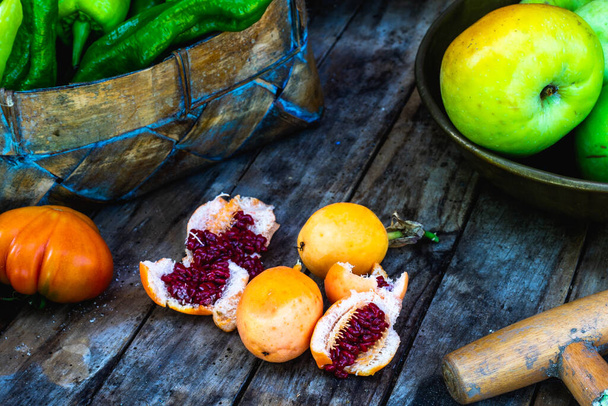 fruto de maracuya aberto ao meio mostrando suas sementes vermelhas em uma mesa de madeira com tomates, maçãs e uma cesta com pimentas - Foto, Imagem