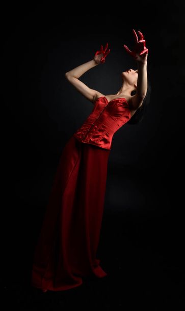 Full length portrait of beautiful young asian woman wearing red corset, covered with bloody paint posing in gothic horror style with creative hand gestures isolated against dark black studio background. - Foto, imagen