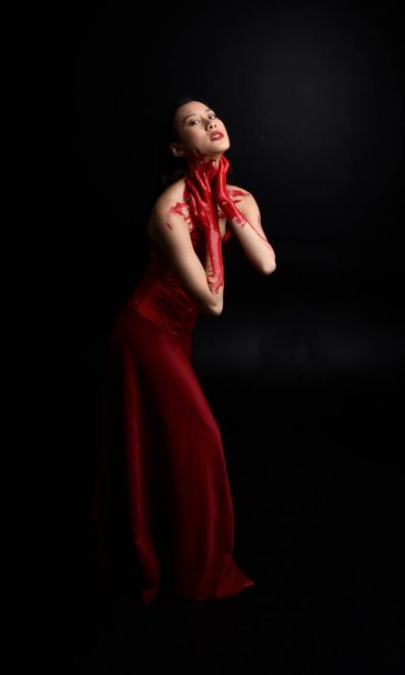 Full length portrait of beautiful young asian woman wearing red corset, covered with bloody paint posing in gothic horror style with creative hand gestures isolated against dark black studio background. - Photo, image