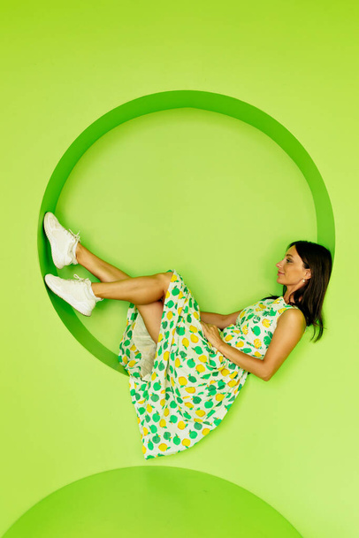 Charmant mooi meisje met donker haar dragen heldere zomer jurk liggend en poseren over geïsoleerde groene achtergrond. Hoge kwaliteit foto - Foto, afbeelding