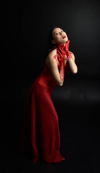 Full length portrait of beautiful young asian woman wearing red corset, covered with bloody paint posing in gothic horror style with creative hand gestures isolated against dark black studio background. - Фото, изображение