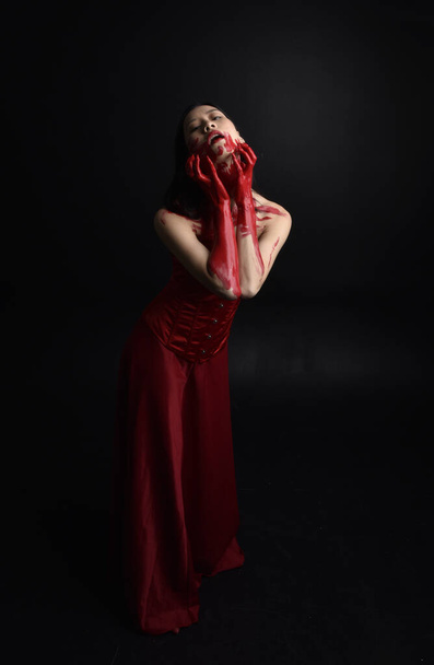 Retrato de larga duración de una hermosa joven asiática vestida con corsé rojo, cubierta con pintura sangrienta posando en estilo de terror gótico con gestos creativos de mano aislados sobre fondo negro oscuro del estudio. - Foto, Imagen