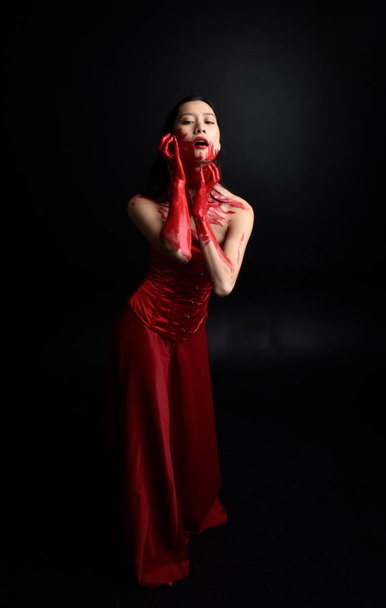 Portret pięknej młodej Azjatki w czerwonym gorsecie, pokrytej krwawą farbą pozującą w gotyckim stylu horroru z kreatywnymi gestami dłoni wyizolowanymi na ciemnym czarnym tle studia. - Zdjęcie, obraz