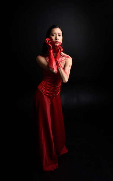 Retrato de larga duración de una hermosa joven asiática vestida con corsé rojo, cubierta con pintura sangrienta posando en estilo de terror gótico con gestos creativos de mano aislados sobre fondo negro oscuro del estudio. - Foto, imagen