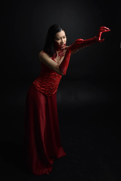 Full length portret van mooie jonge aziatische vrouw dragen rood korset, bedekt met bloederige verf poseren in gotische horror stijl met creatieve hand gebaren geïsoleerd tegen donker zwart studio achtergrond. - Foto, afbeelding