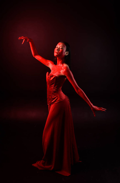 Full length portrait of beautiful young asian woman wearing red corset, covered with bloody paint posing in gothic horror style with creative hand gestures isolated against dark black studio background. - Valokuva, kuva