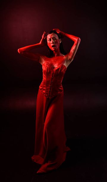 Full length portrait of beautiful young asian woman wearing red corset, covered with bloody paint posing in gothic horror style with creative hand gestures isolated against dark black studio background. - Zdjęcie, obraz