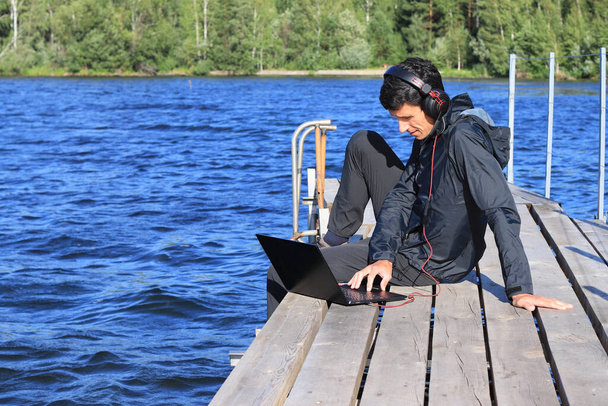 O jovem trabalha remotamente. Um homem está sentado em um cais de madeira junto à água com fones de ouvido e um laptop. Homem a trabalhar remotamente na natureza. Conceito de trabalho remoto, freelance. - Foto, Imagem