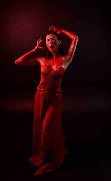 Ganztägiges Porträt einer schönen jungen asiatischen Frau, die ein rotes Korsett trägt, mit blutiger Farbe bedeckt und im gotischen Horror-Stil posiert, mit kreativen Handgesten isoliert vor dunkelschwarzem Studiohintergrund. - Foto, Bild