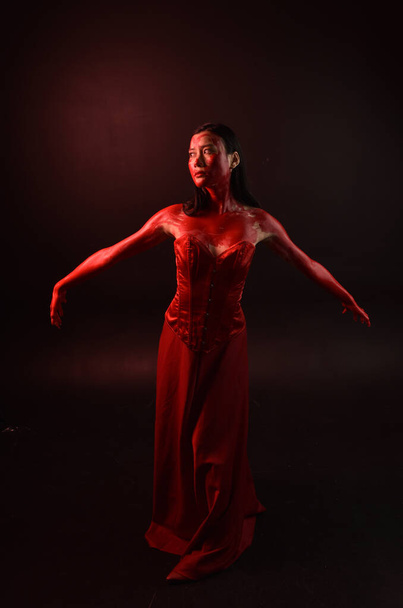 Полный портрет красивой молодой азиатской женщины в красном корсете, покрытый окровавленной краской, позирующей в готическом стиле ужасов, с творческими жестикуляциями рук, изолированными на темном черном фоне студии. - Фото, изображение