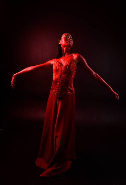 Full length portret van mooie jonge aziatische vrouw dragen rood korset, bedekt met bloederige verf poseren in gotische horror stijl met creatieve hand gebaren geïsoleerd tegen donker zwart studio achtergrond. - Foto, afbeelding